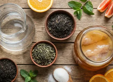 Una fotografía cenital muestra los ingredientes para hacer kombucha sobre una mesa de madera rústica. En el centro hay un frasco de vidrio con una bebida fermentada y un SCOBY flotando. Alrededor se disponen una jarra con agua, tres cuencos con té negro y verde suelto, una cuchara con azúcar blanca, y rodajas de naranja, pomelo y limón, junto con hojas de menta.